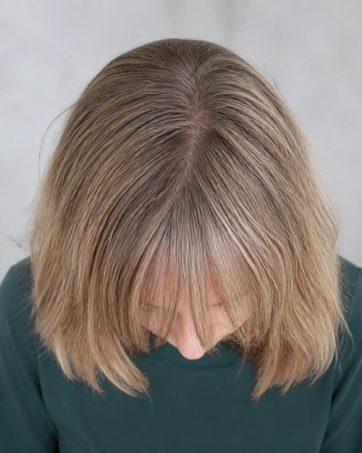 Overhead view of a mature woman with dark blonde hair, head tilted forward, showing long undone bangs extending past the brow area in intentionally imperfect separated pieces