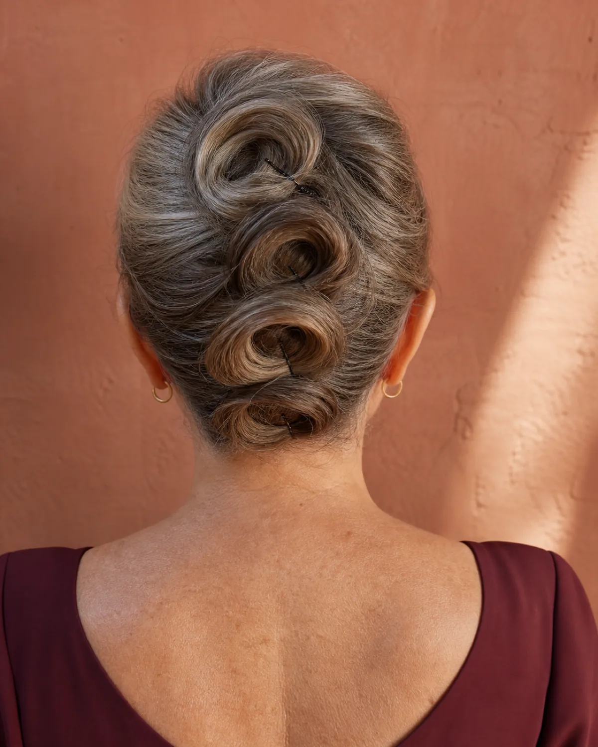 Back view from slightly above of a woman with dark hair arranged in three distinct pinned loops at the back of the head, each loop holding its shape as a separate element, warm directional light from the upper left