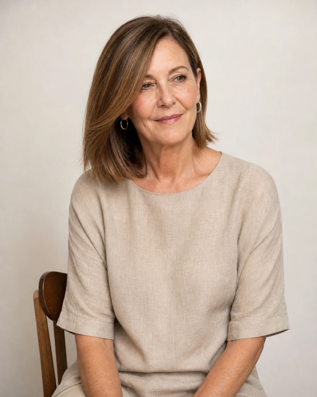 A woman in her early 60s with warm brown hair cut to above-the-shoulder length, seated on a wooden chair, the clean line ending two inches above the shoulder seam