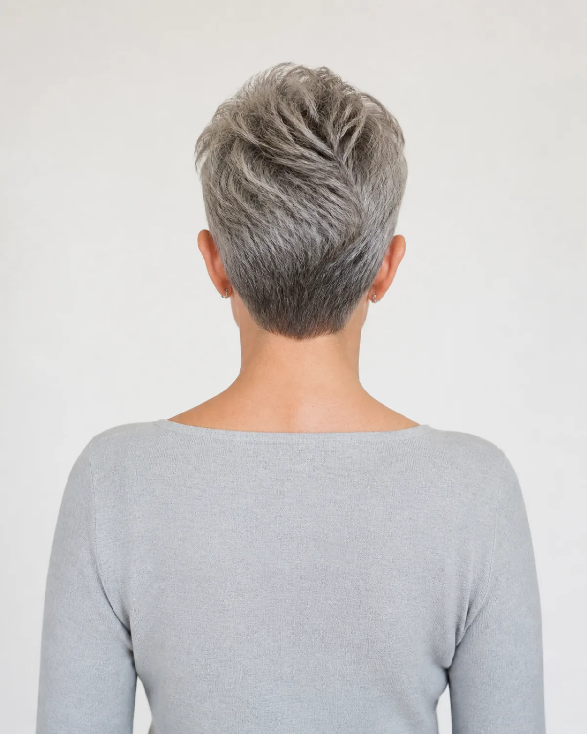 Back view of a woman with warm grey hair in a very short crop, the crown volume standing clearly higher than the close-cropped sides, off-white background