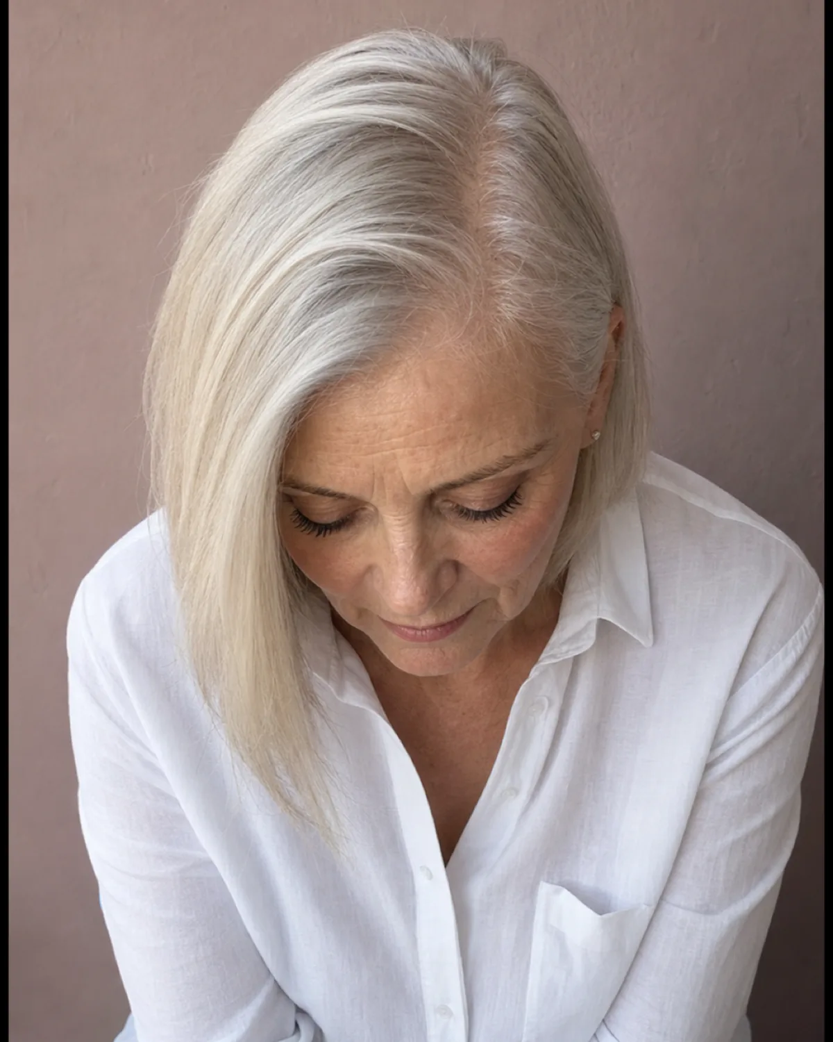 A woman with fine platinum-blonde hair at chin length shot from slightly above, a deep side parting displacing most of the hair to her left shoulder while the right side shows thinner coverage