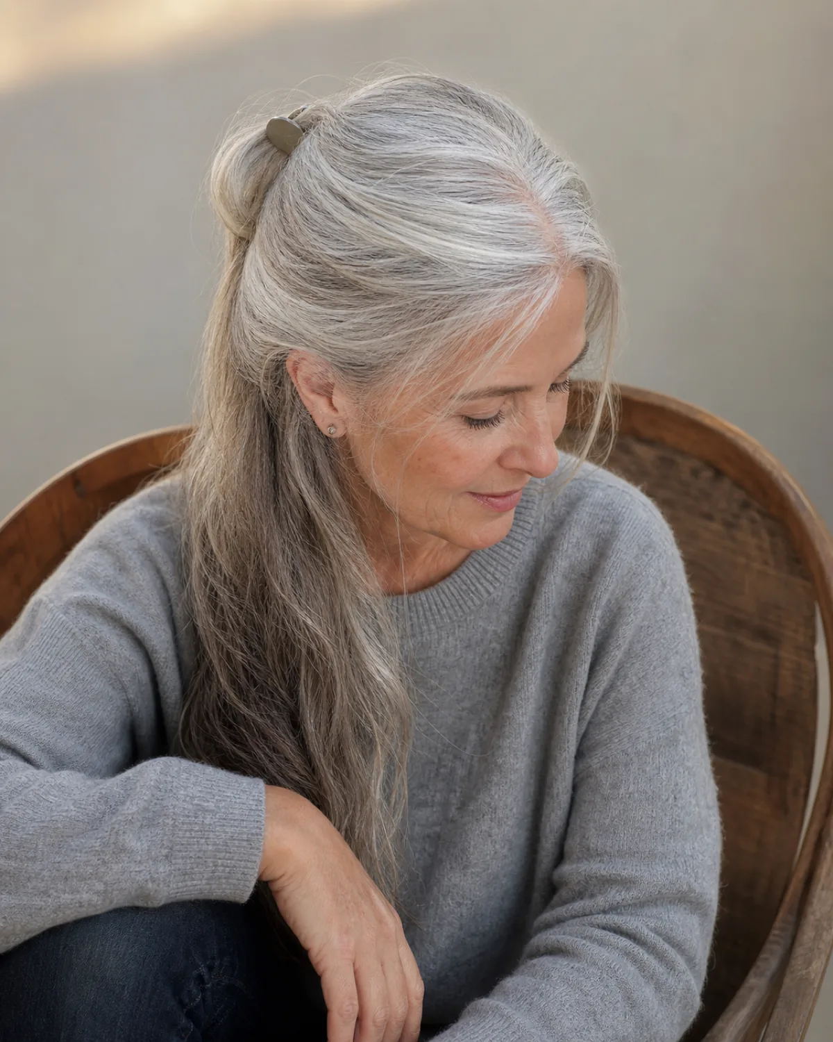 A woman in her early 60s seated in a wooden chair, shot from slightly above, silver-grey hair in a half-up half-down style with the top section gathered at the crown by a matte clip and the bottom half falling forward over one shoulder, wearing a grey cashmere pullover