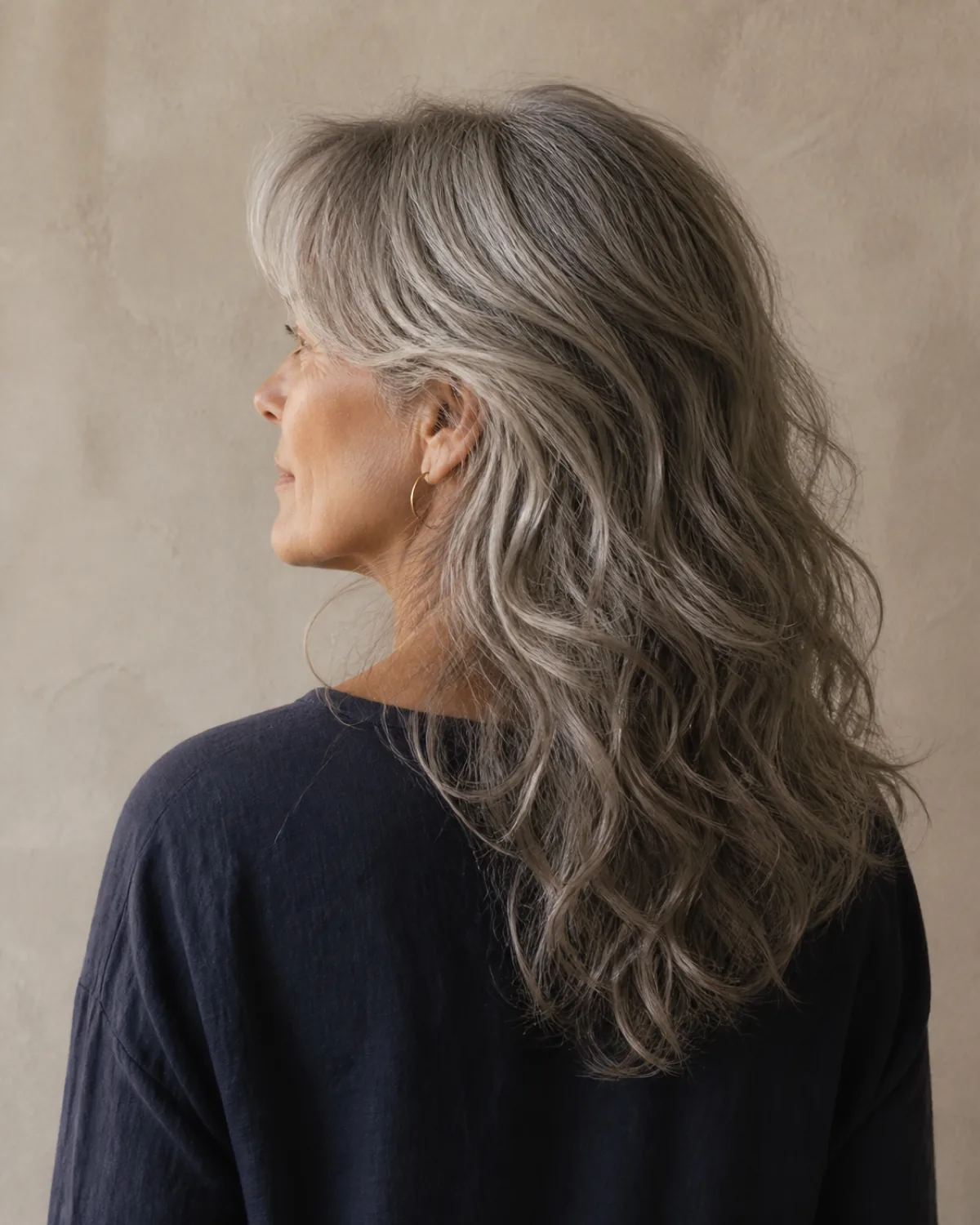 Back-three-quarter view of a woman in her late 50s with wavy salt-and-pepper grey hair in a long shag, curtain bangs sweeping to one side, visible disconnected layers creating movement, wearing a navy linen top against a stone wall
