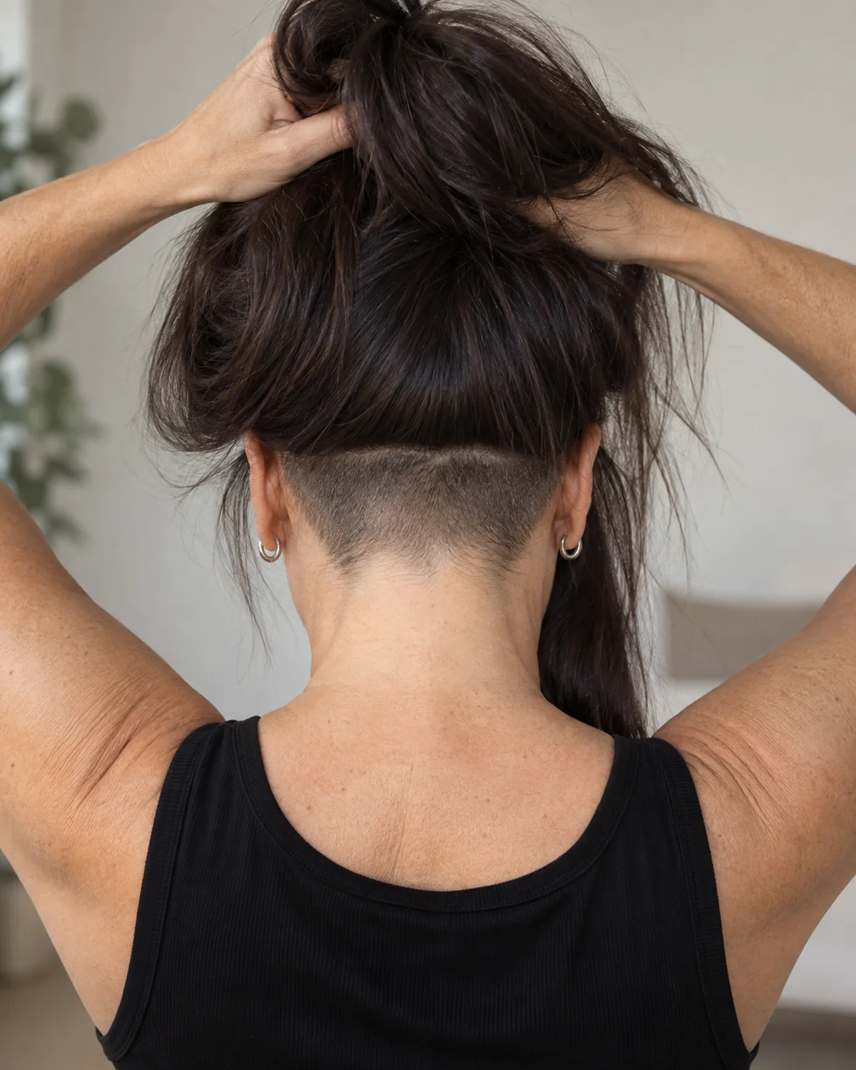Mature woman from behind lifting her thick dark hair to show a closely clippered undercut at the nape, black sleeveless top