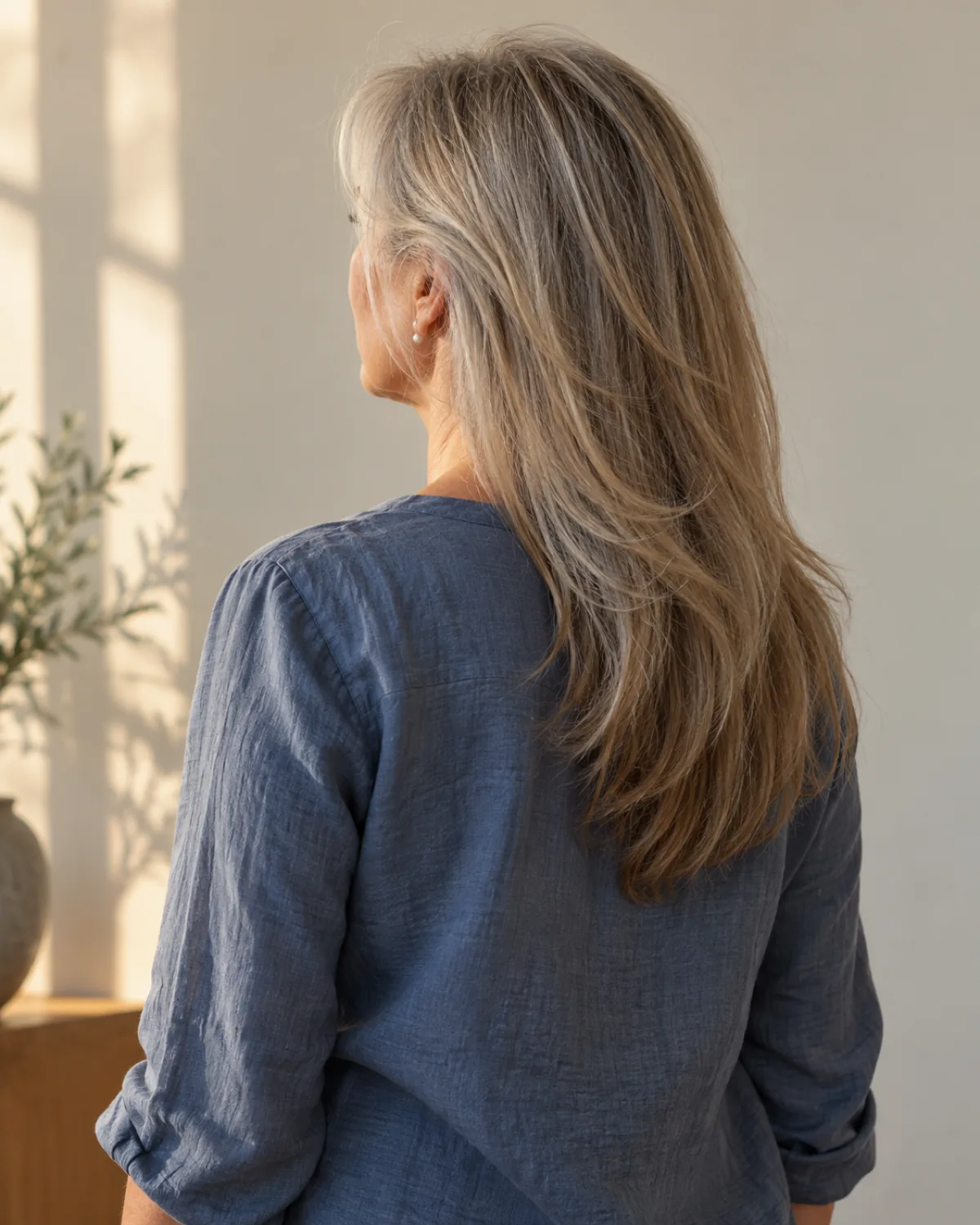 Back-three-quarter view of mature woman with fine layered hair falling to mid-back, shortest layer starting below the jawline, slate blue linen blouse, warm afternoon light