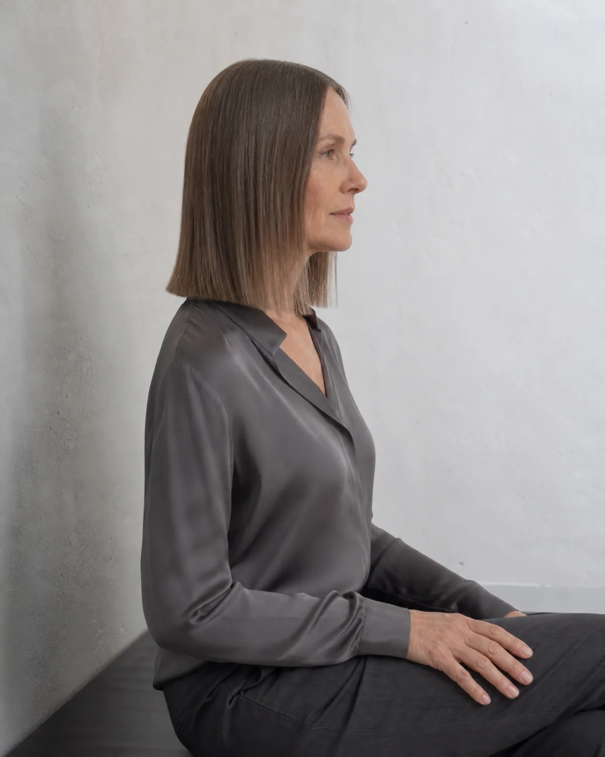 A mature woman with fine cool-brown hair in a blunt lob with a precise centre parting, viewed in a seated side profile with one hand resting on her lap, the geometric perimeter falling to the collarbone against a white plaster wall