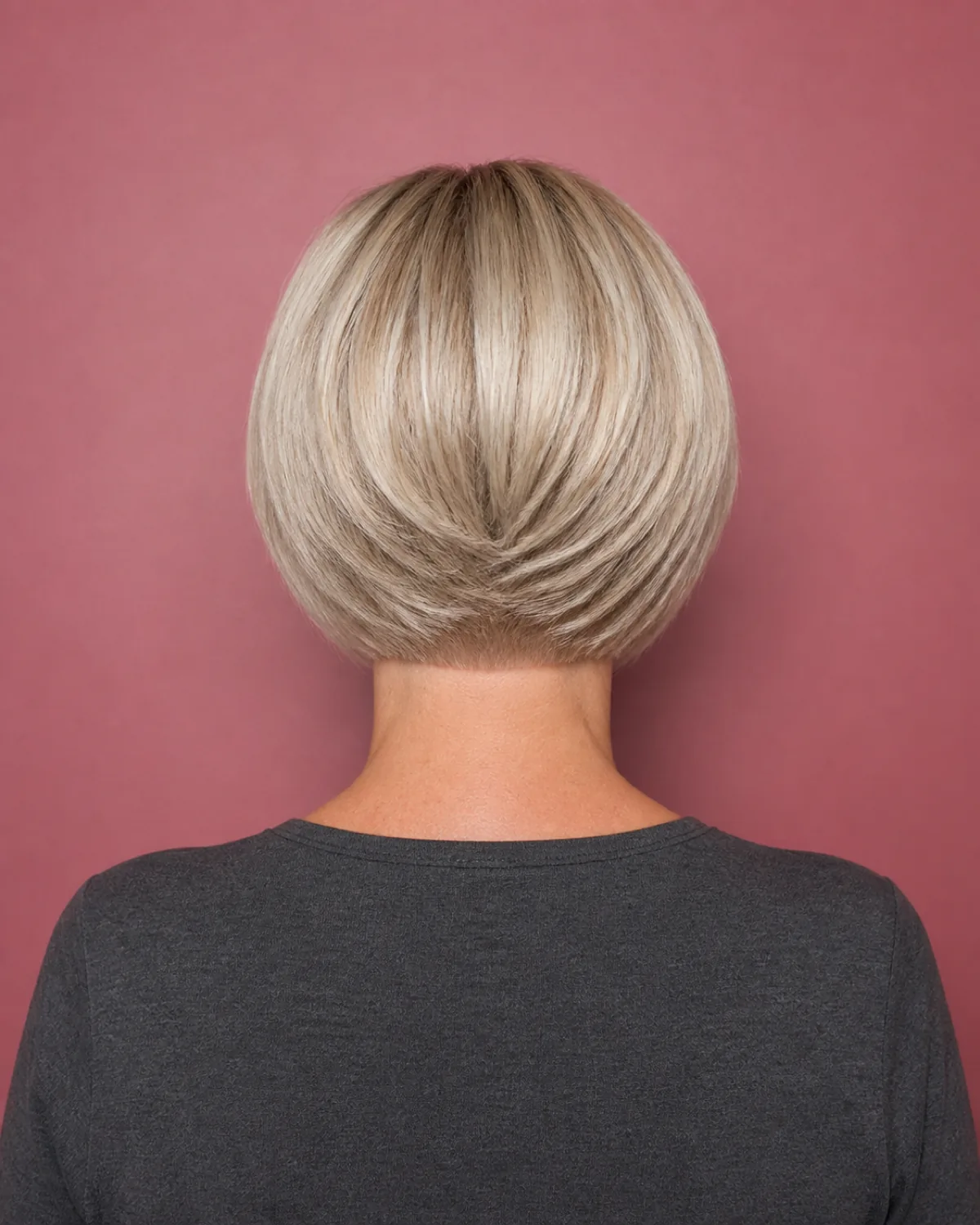 A mature woman with fine cool-blonde hair in a chin-length graduated bob viewed from directly behind, the graduation at the nape clearly visible with each layer stepping inward to create a wedge shape against a muted rose-toned wall