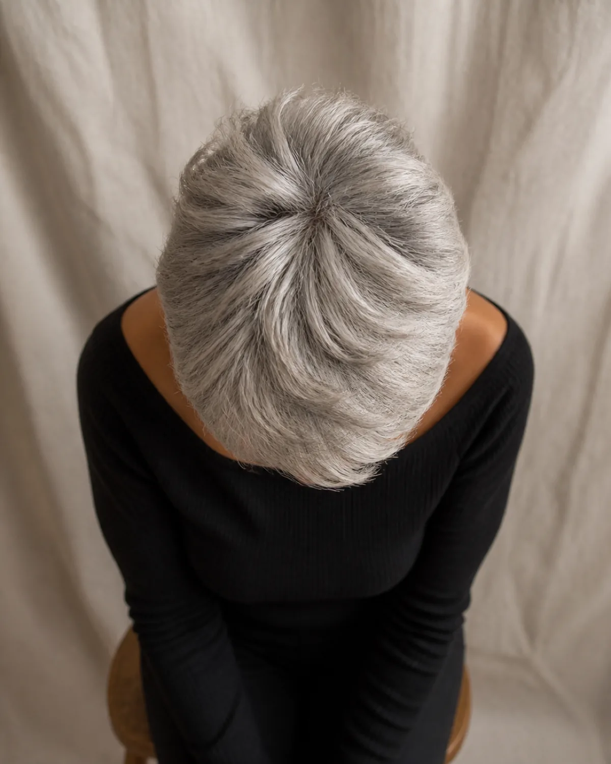Overhead view of a mature woman with fine silver hair in a textured pixie cut, the crown carrying visible length and lift while the sides are closely cropped, warm studio light picking out individual strands