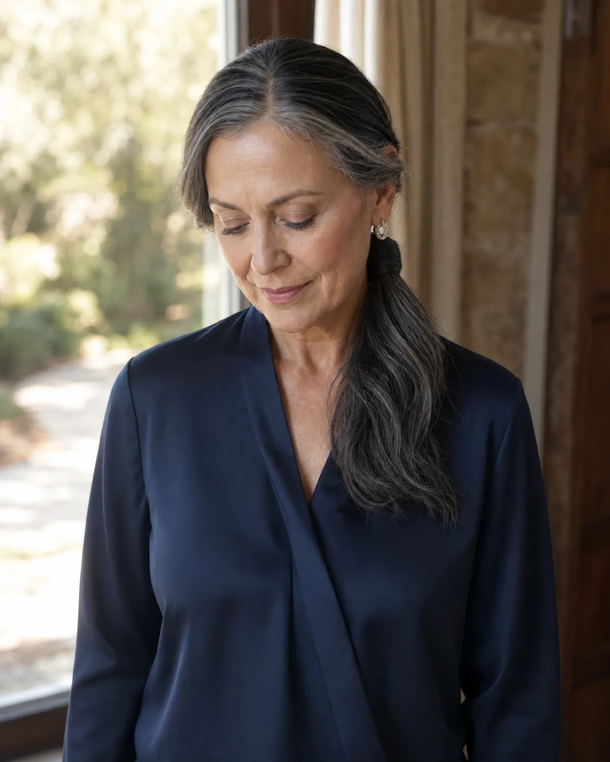 Front view of a woman with dark silver-streaked hair in a wrapped low ponytail, eyes downcast, a section of hair wrapped around the elastic at the base, wearing a navy silk blouse, garden behind