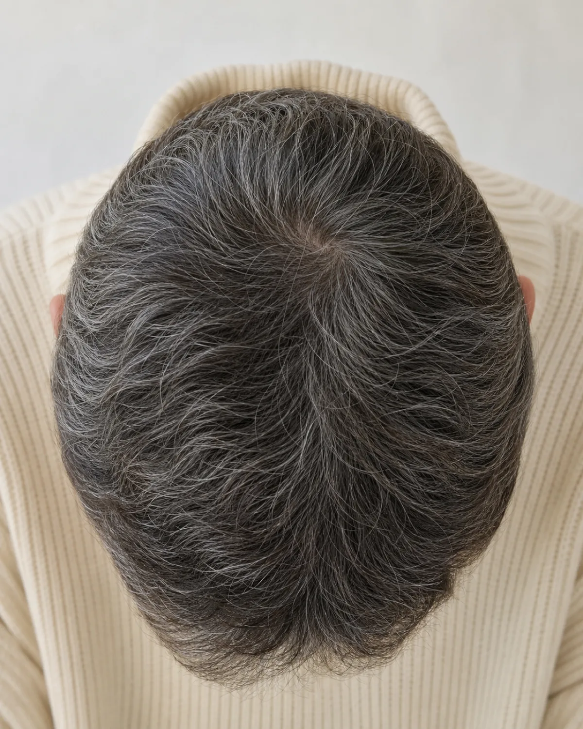 A top-down shot of a woman's crown showing salt-and-pepper hair in a textured crop, dark brown and silver-grey strands naturally interspersed, wearing a cream turtleneck