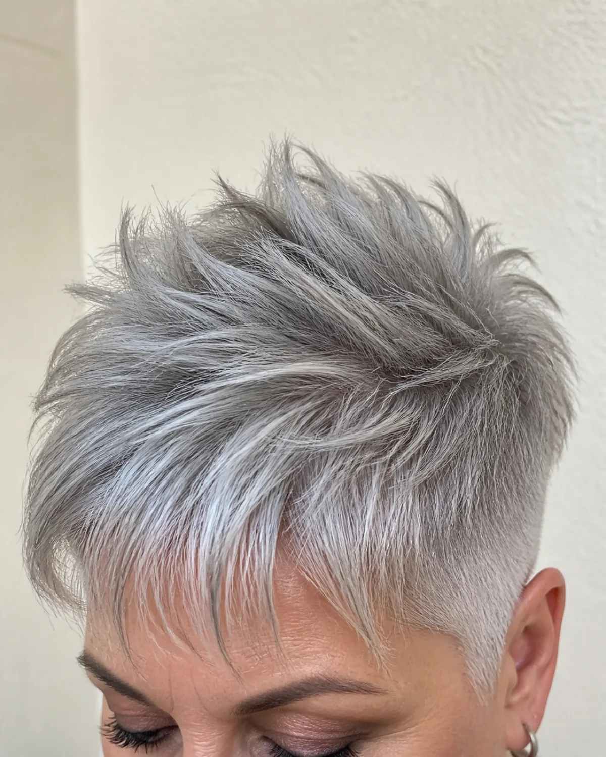 Close crop on the crown and fringe of a woman over 50, silver-grey pixie with tapered short sides and dramatically more length and lift at the crown, soft daylight from above, cream wall