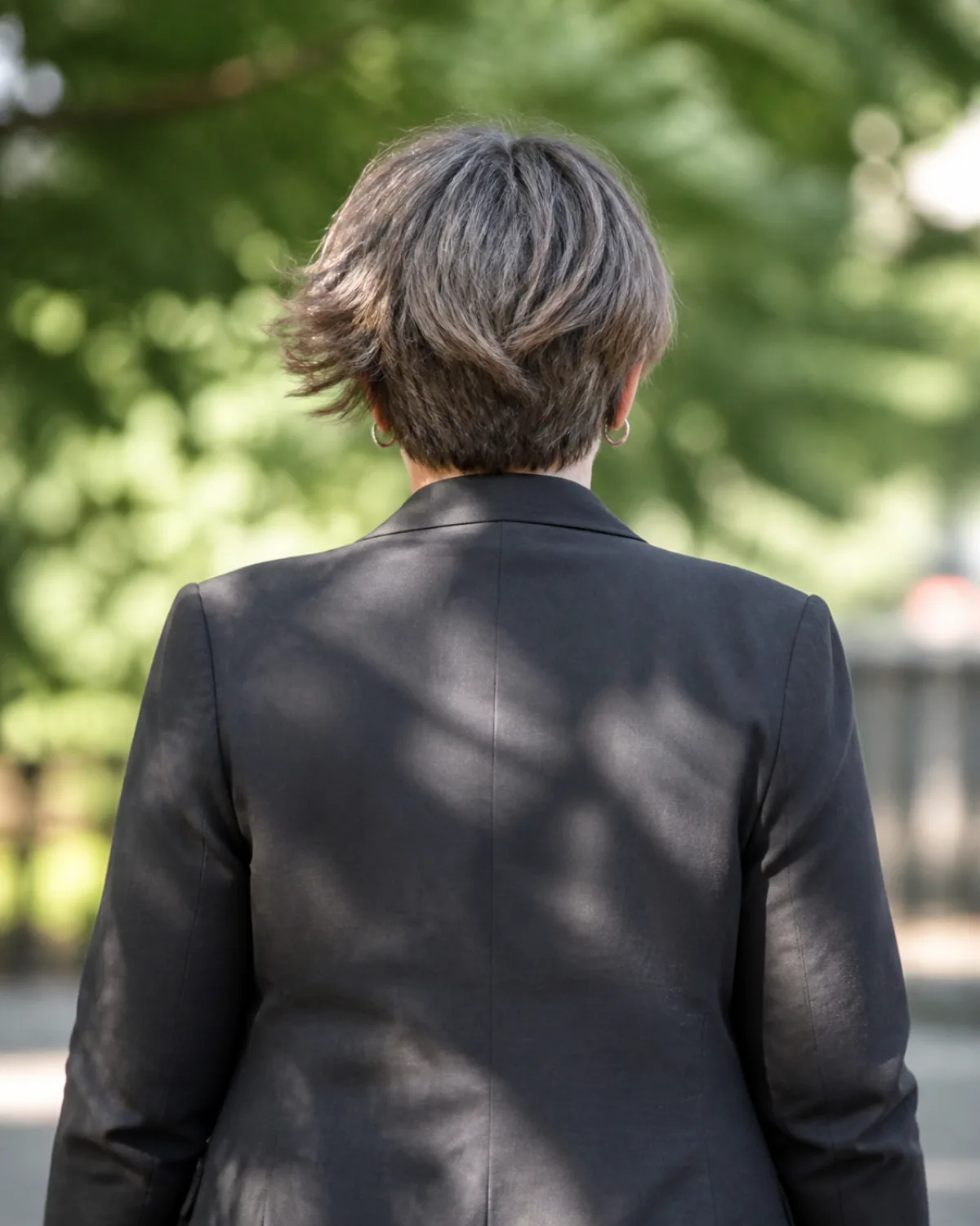 A woman over 50 with a fuller build walking away from the camera, textured pixie bob with choppy ash-brown and silver pieces between the ears and jaw, charcoal blazer, dappled light with blurred outdoor green