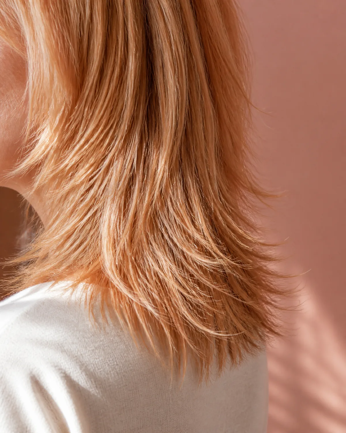 Close crop of fine strawberry-blonde hair showing soft feathered razored ends tapering to individual strands, dusty-rose wall