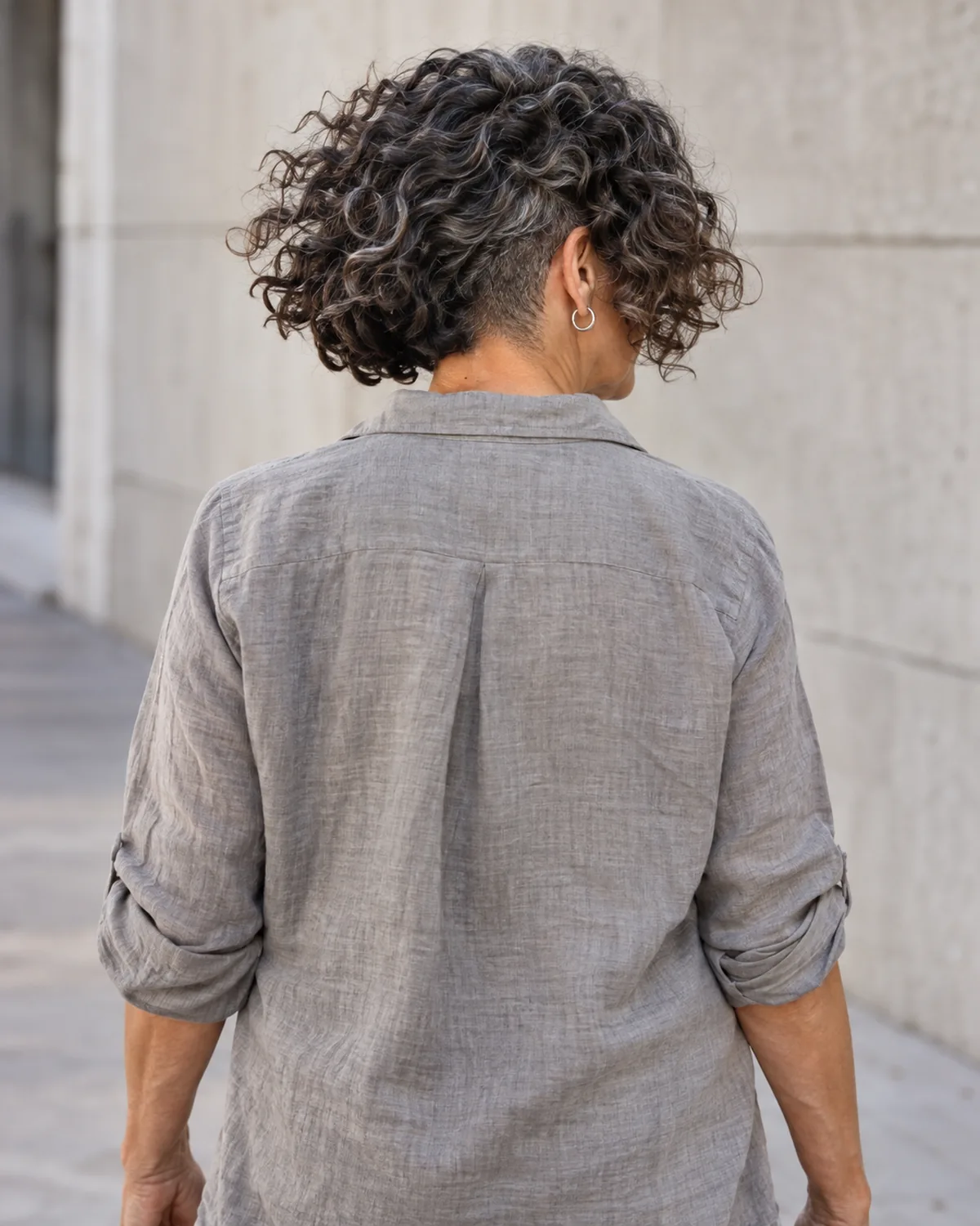 Rear view of mature woman walking, thick dark curly bob with silver threads, undercut visible where curls part with movement