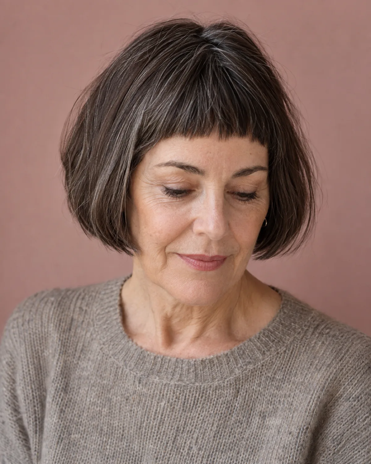 Front-facing view with eyes downcast of a mature woman with thick dark brown hair with scattered silver in a French bob with blunt bangs above the eyebrows