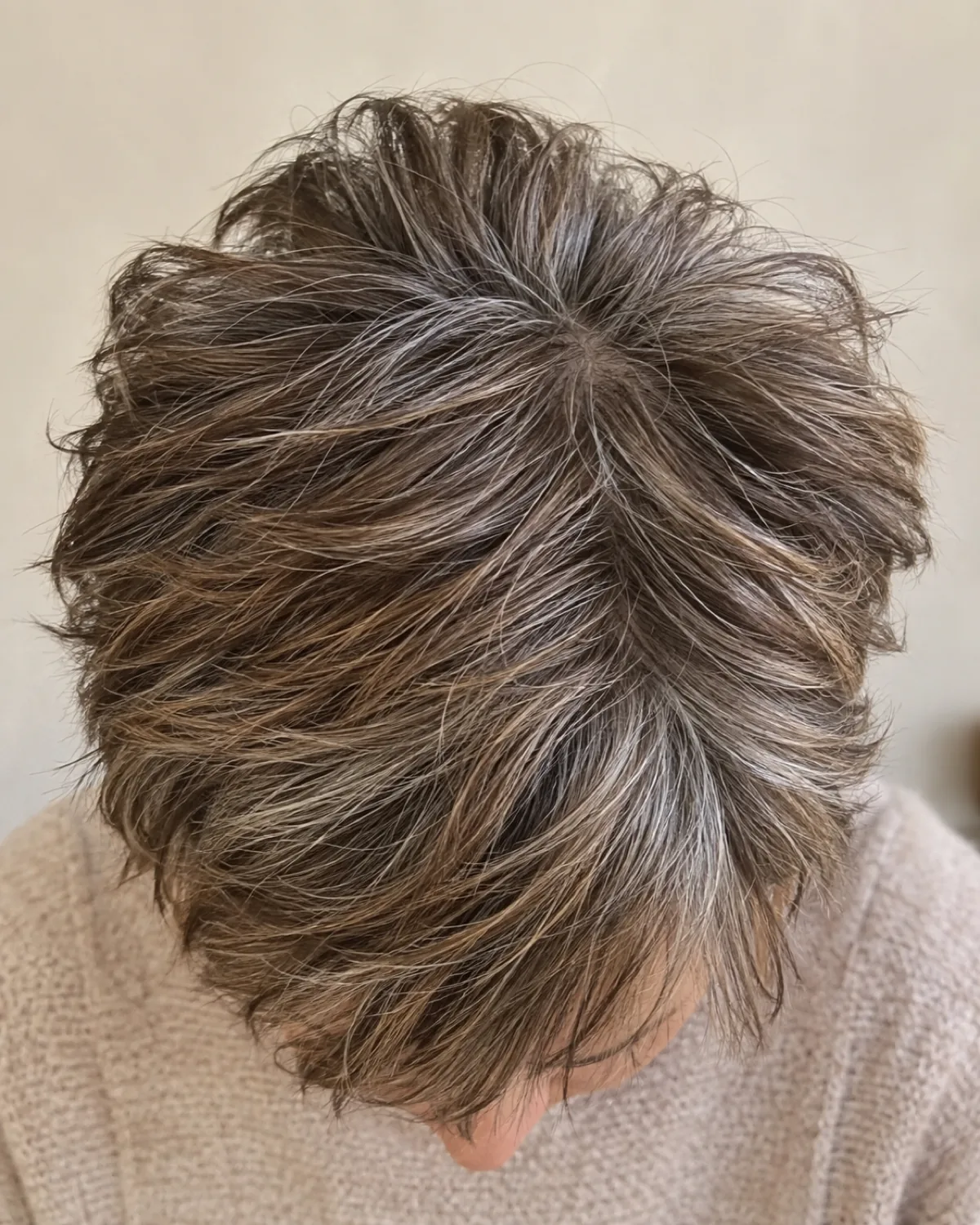 Top-of-head view of a mature woman with thick warm brown hair with heavy grey showing a shaggy pixie with visibly razored choppy ends and natural texture