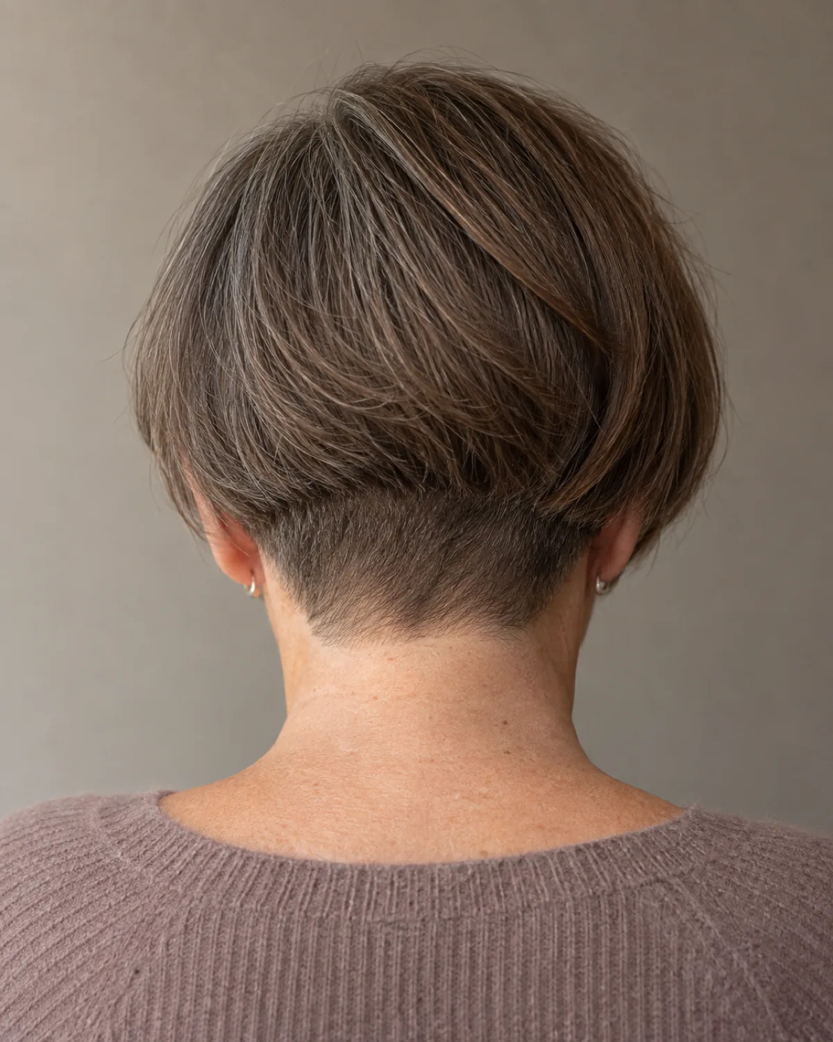 Back view of a mature woman with thick medium brown hair showing an undercut bob with the nape section clippered short underneath and the top bob layer falling over it