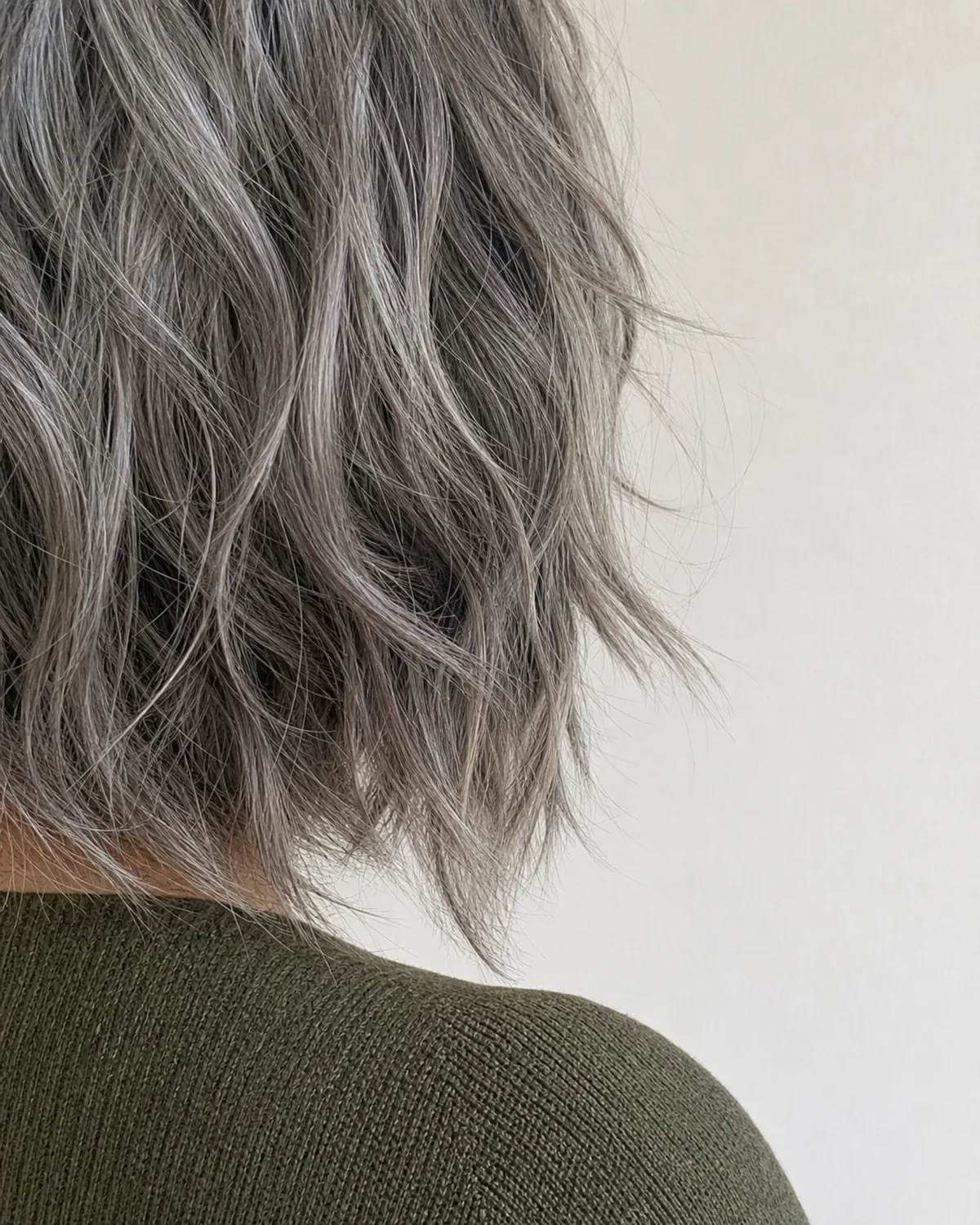 A close crop of the wavy silver ends of a jaw length shag, the short choppy layers finishing at different lengths so the ends separate into distinct pieces above a moss green knit shoulder