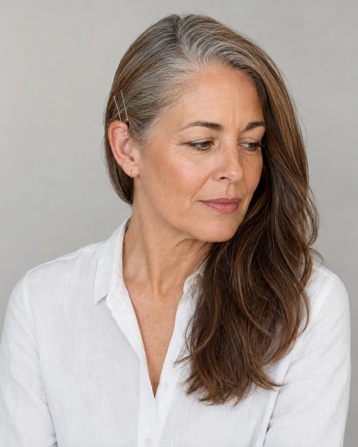 Three-quarter view of a mature woman with warm brown hair and grey roots swept to one side over the shoulder, bobby pins visible behind the opposite ear