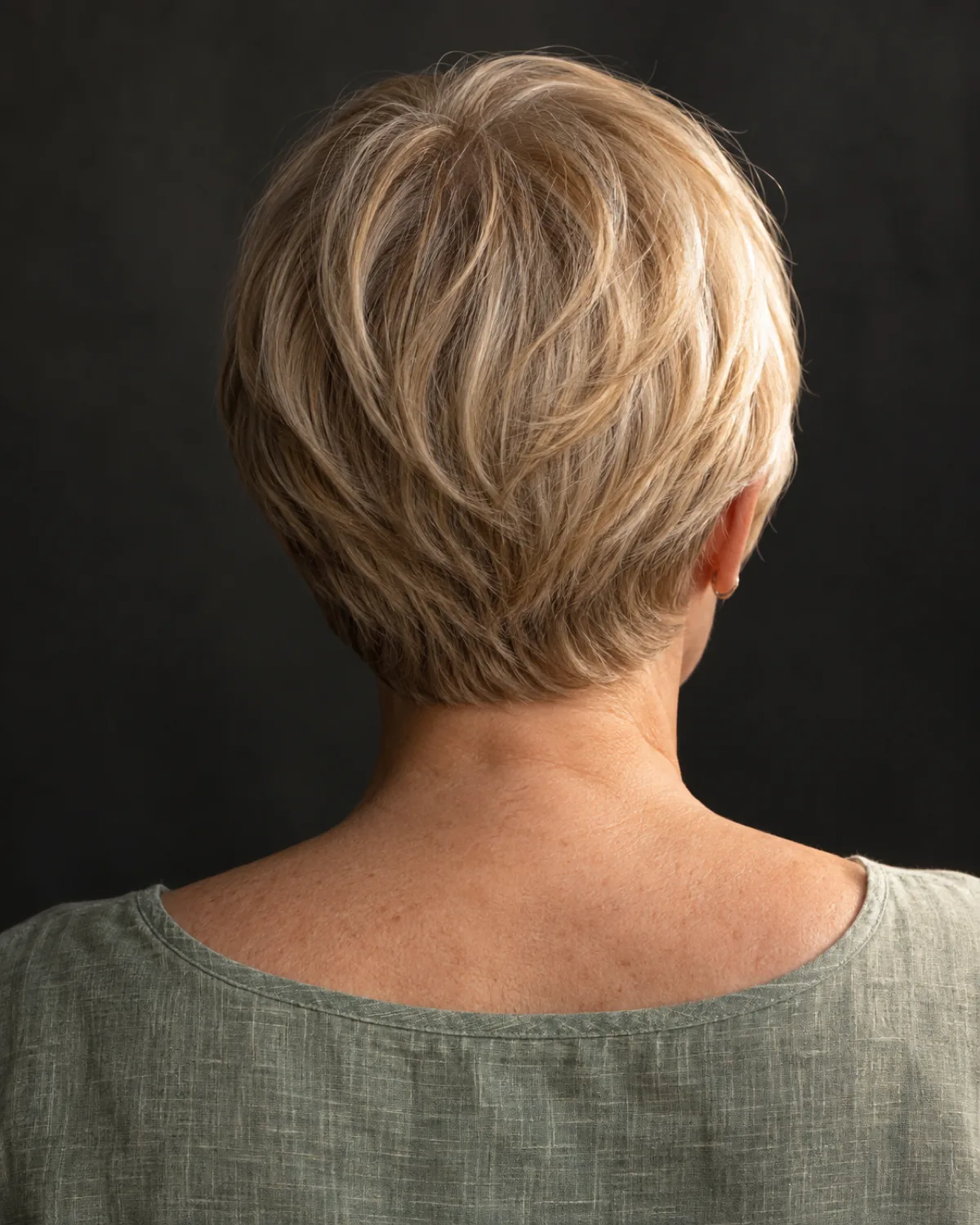 Back three-quarter view of mature woman with soft rounded crop in warm blonde, head-hugging shape, dark charcoal backdrop