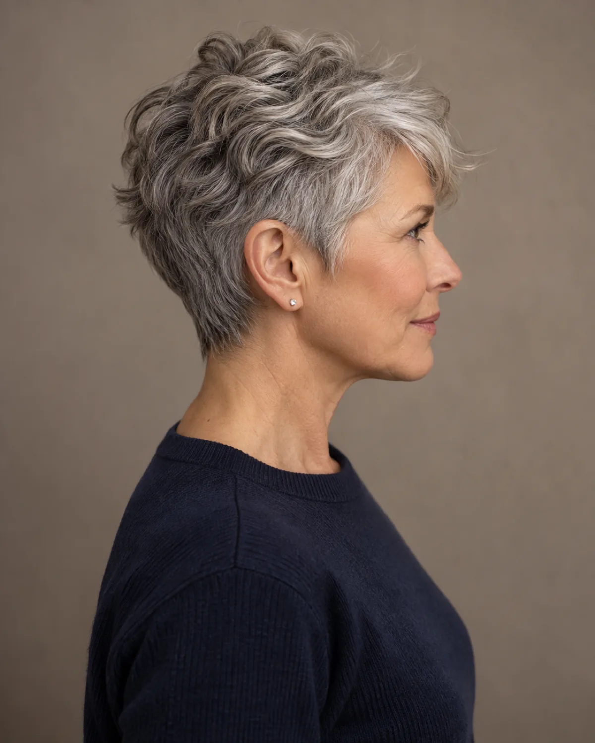 Woman over 50 with silver-grey wavy textured pixie cut seen from the side profile, navy crew-neck sweater, warm grey backdrop