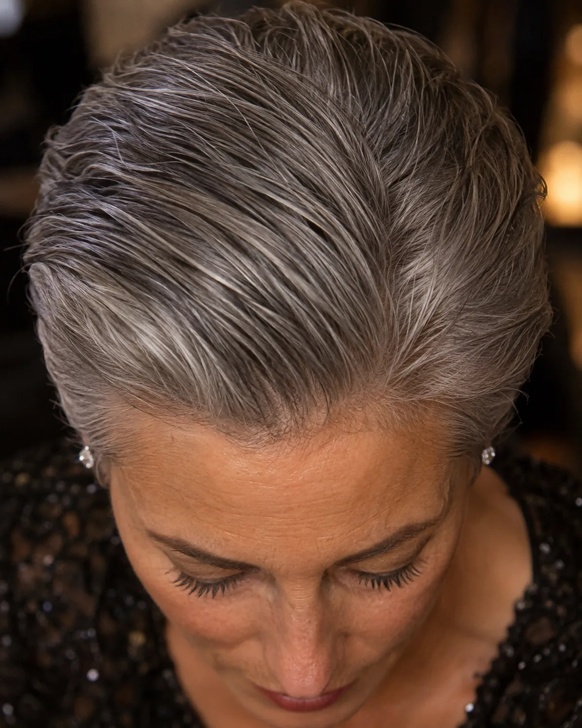 Top-down macro view of a woman's steel-grey cropped hair slicked back with light gel, diamond stud earrings visible